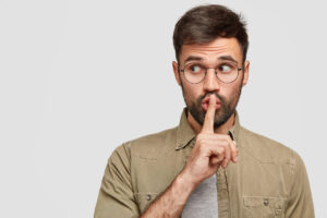 Photo of secret young man makes shush gesture, looks surprisingly aside, notices something strange, has dark stubble, isolated over white background with copy space for your promotional text